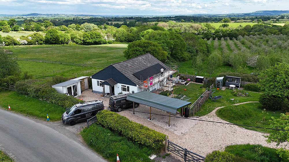 Aerial view of a rural house with scaffolding, two black vans, fields and trees